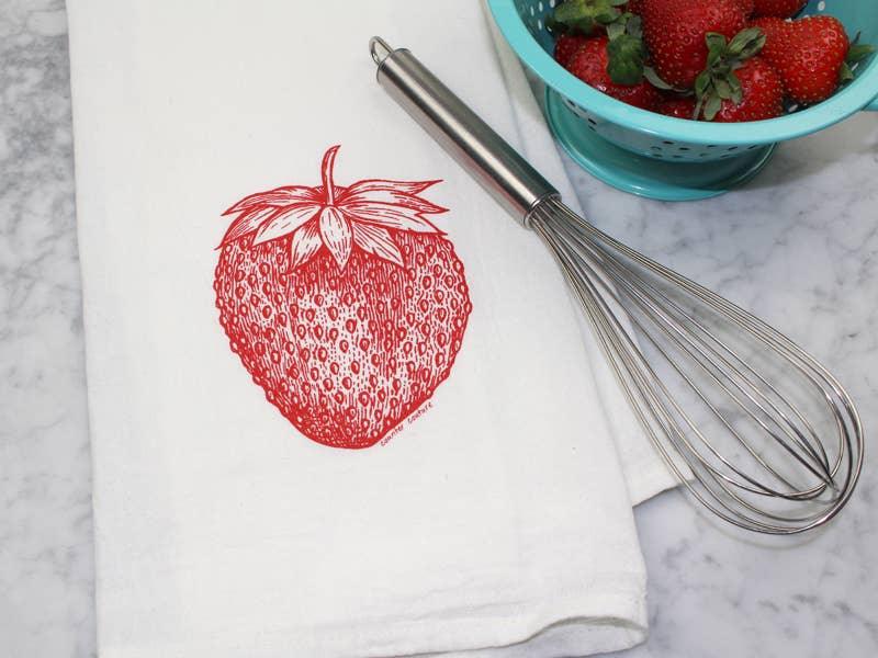 White kitchen towel with red strawberry print, whisk, and fresh strawberries in colander on marble