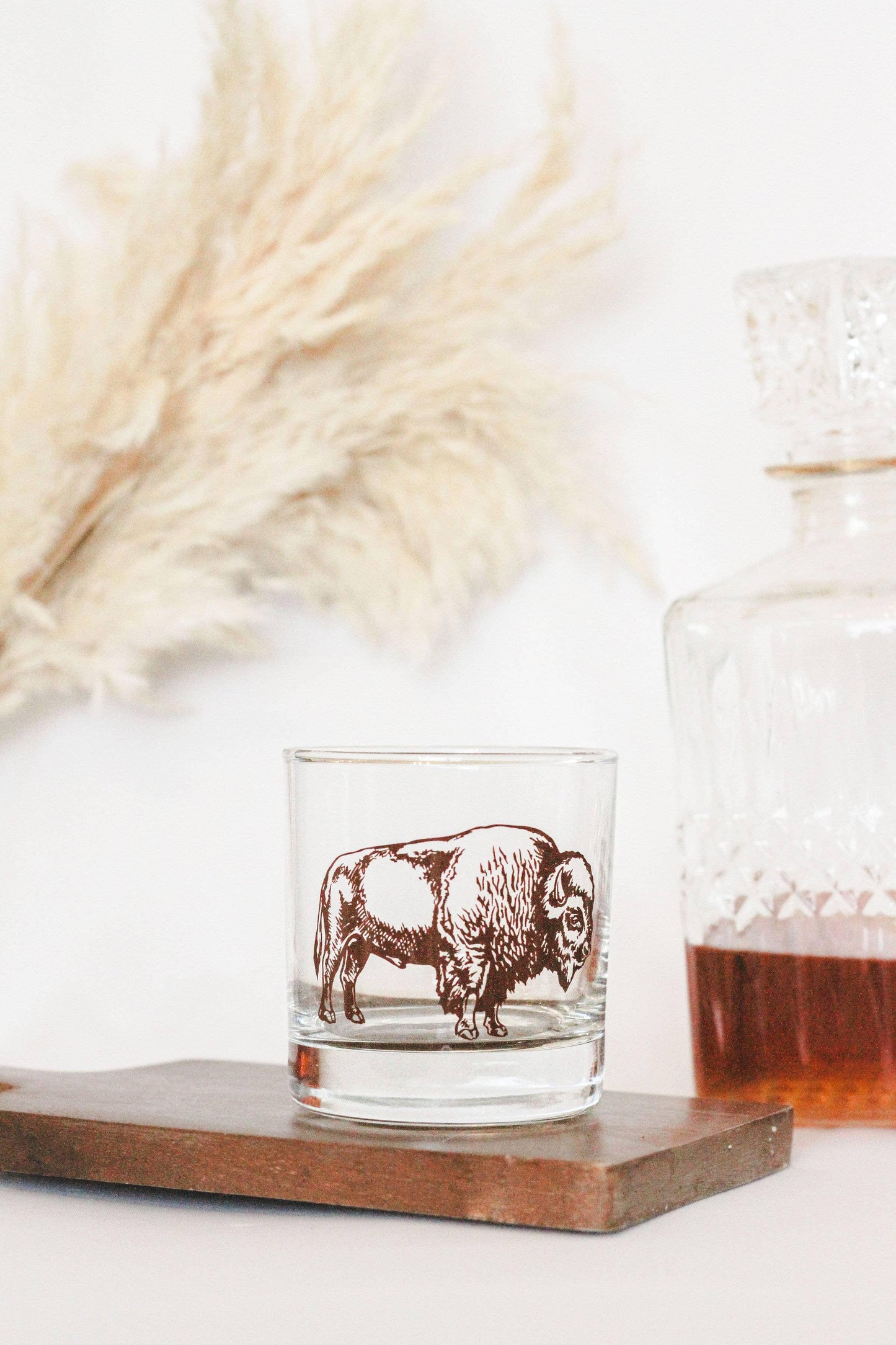 Clear whiskey glass with brown bison design, set on wood tray beside decanter and dried grasses