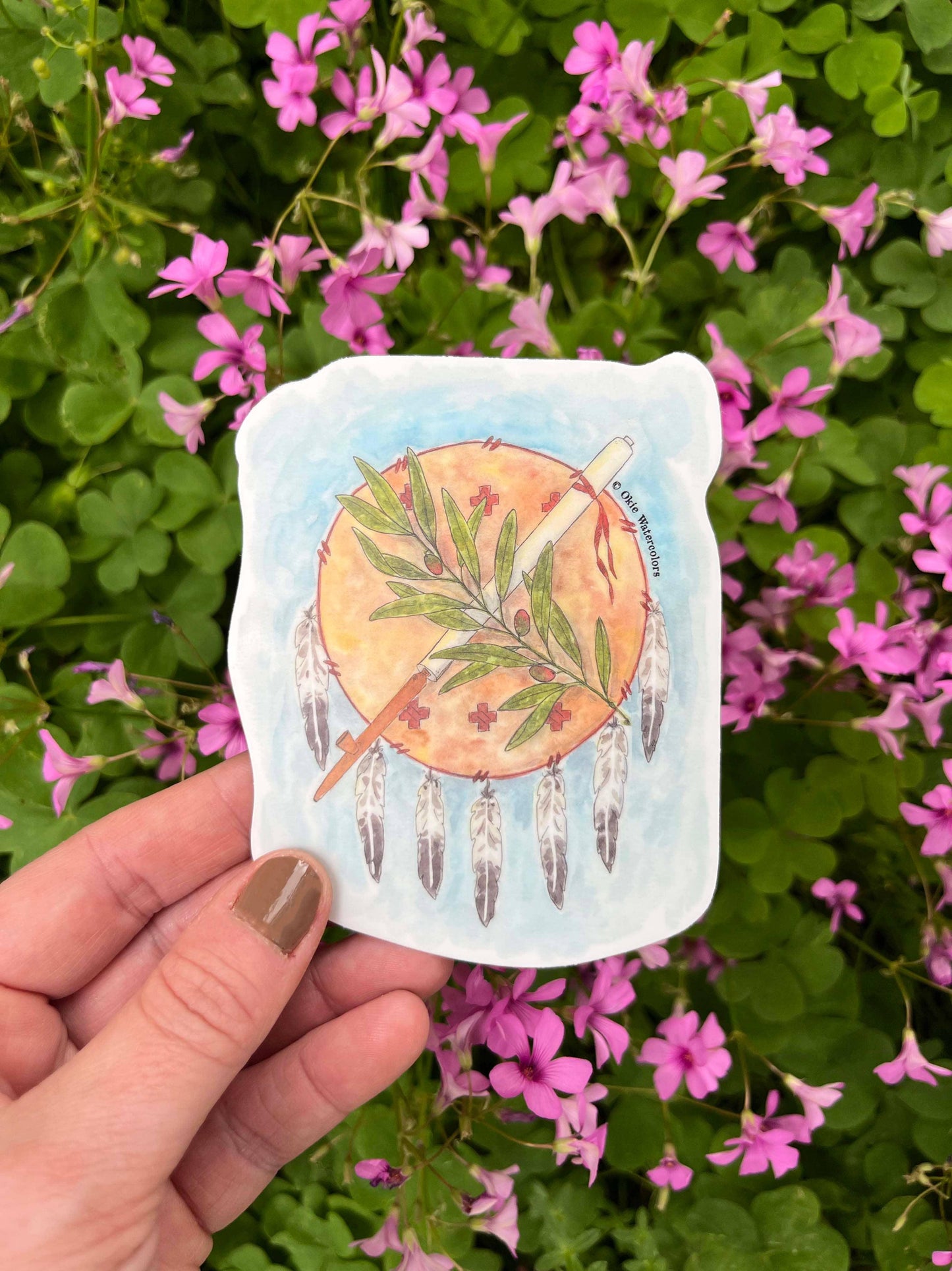 Hand holding Native American dreamcatcher art card with feathers, leaf, and smudge stick, set against pink flowers.