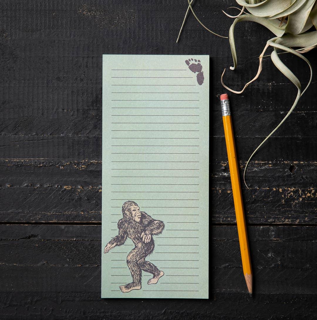 Bigfoot-themed lined notepad with footprint design, pencil, and air plant on dark wood