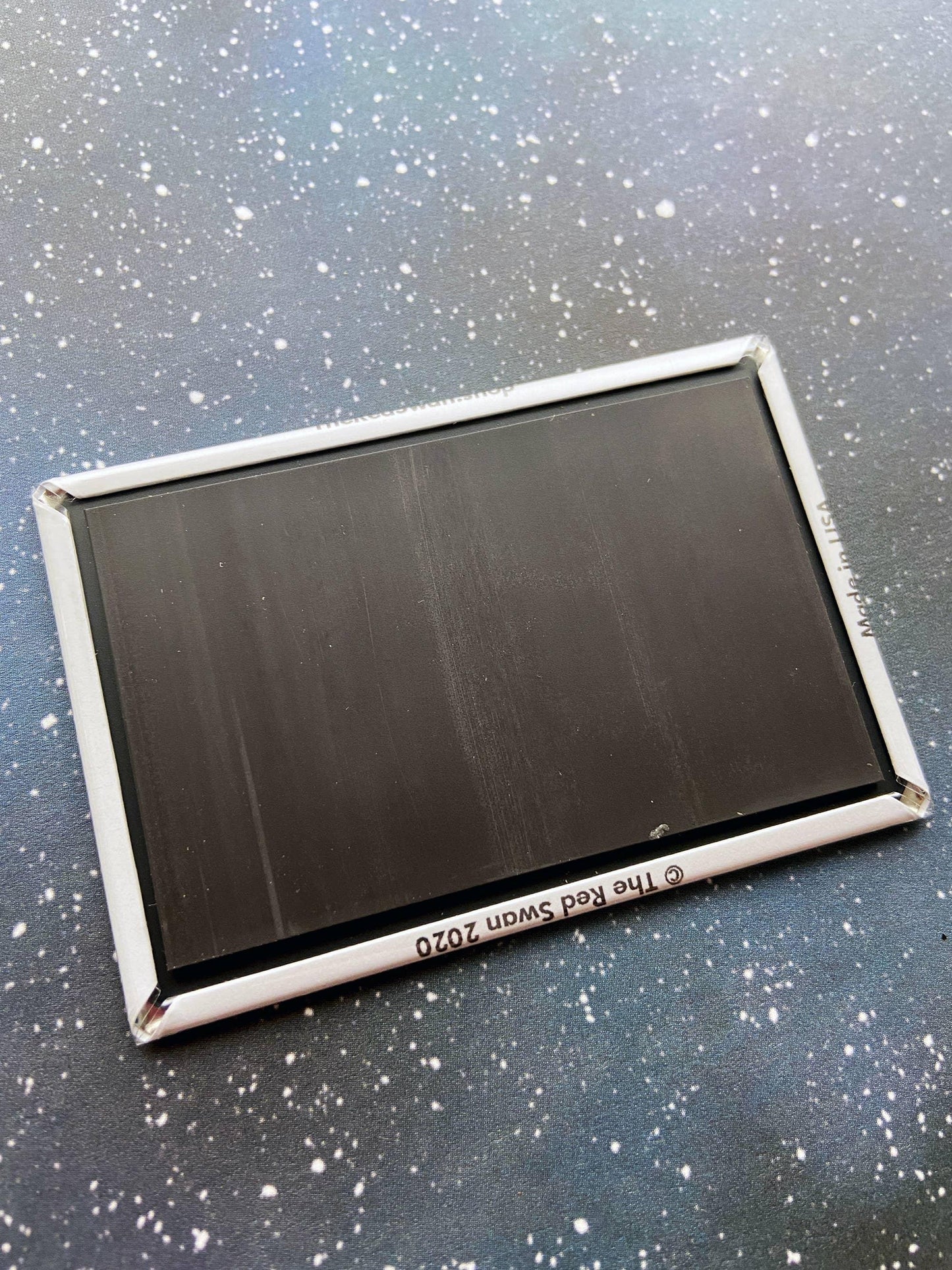 Rectangular black magnet with silver border, © The Red Swan 2020, Made in USA, on speckled surface