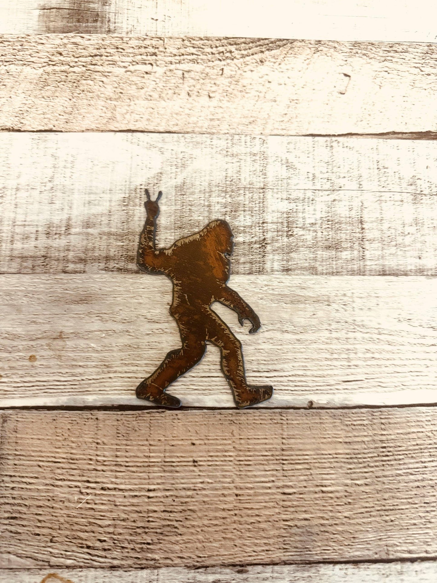 Rustic metal Bigfoot cutout making peace sign on weathered wood, Native art craft supply
