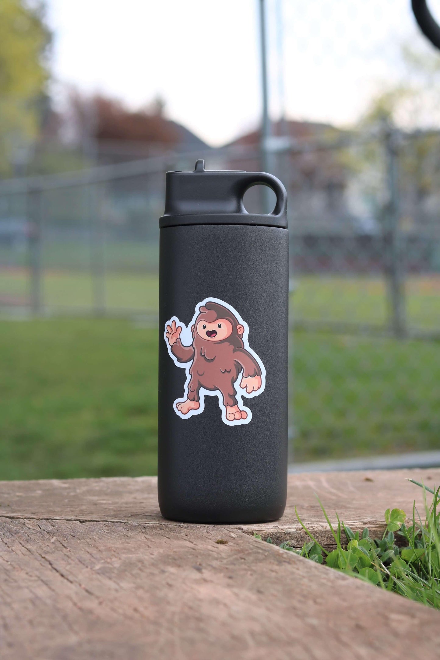 Black water bottle with cute cartoon Bigfoot sticker, sitting on wooden table outdoors