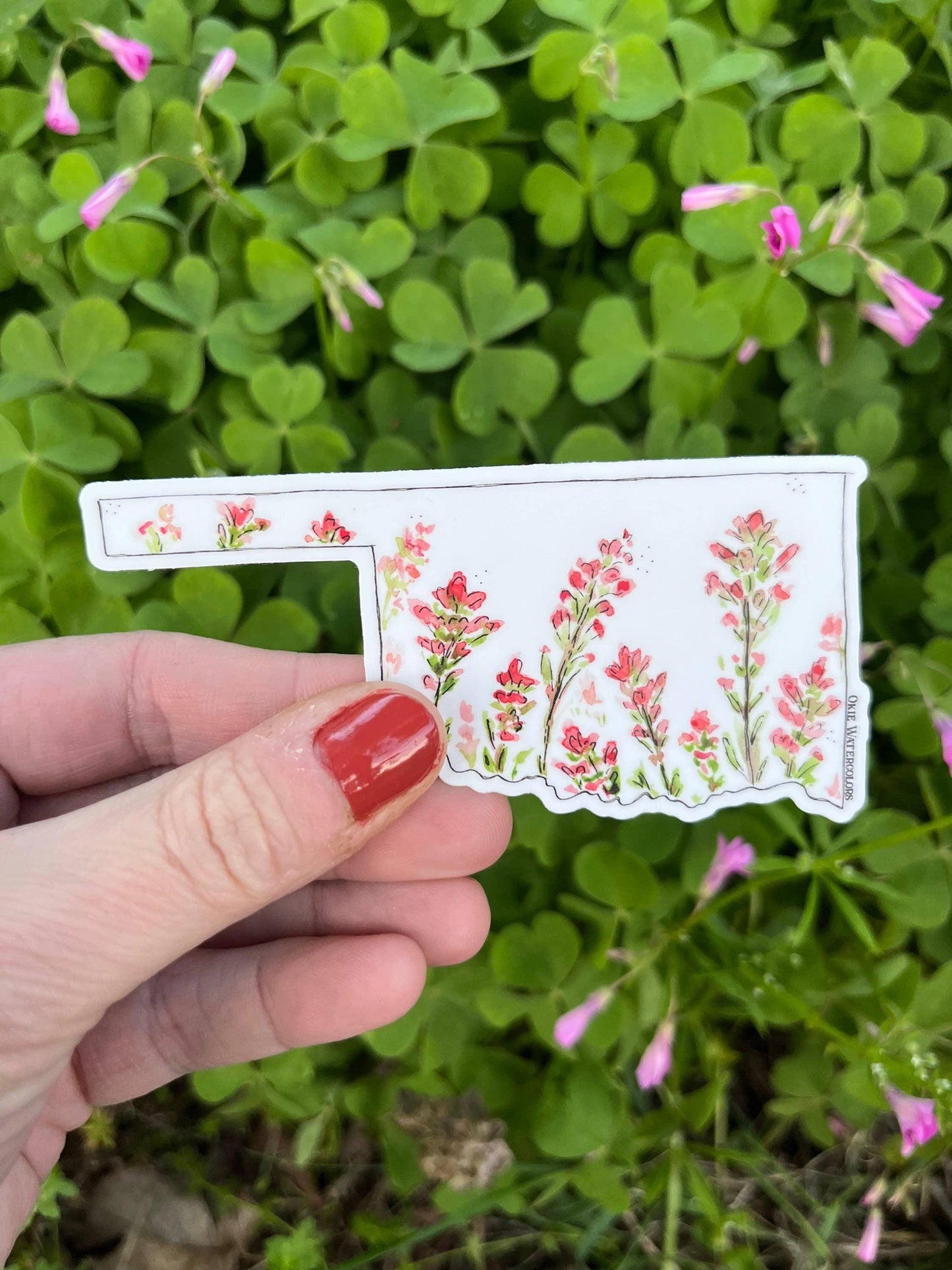Hand holding Oklahoma-shaped sticker with watercolor wildflowers, set against green clover and pink blooms