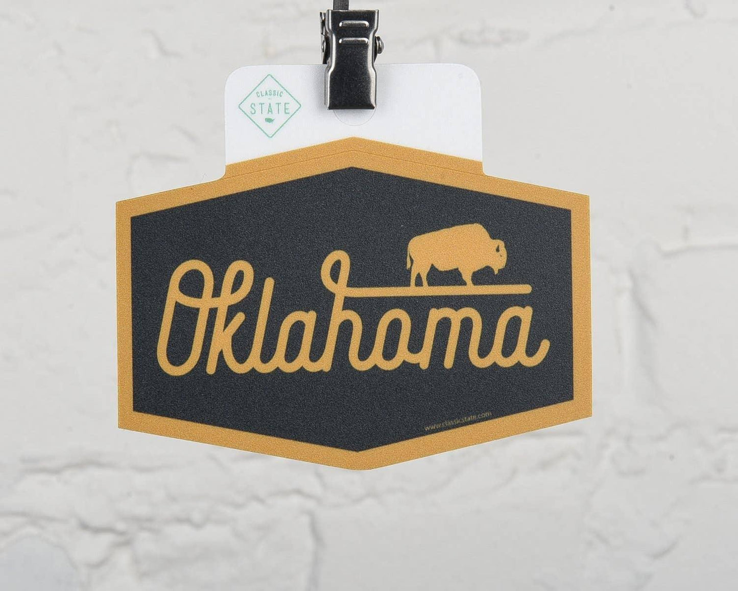 Oklahoma state sticker with bison design, gold and black, hanging on clip against white brick wall
