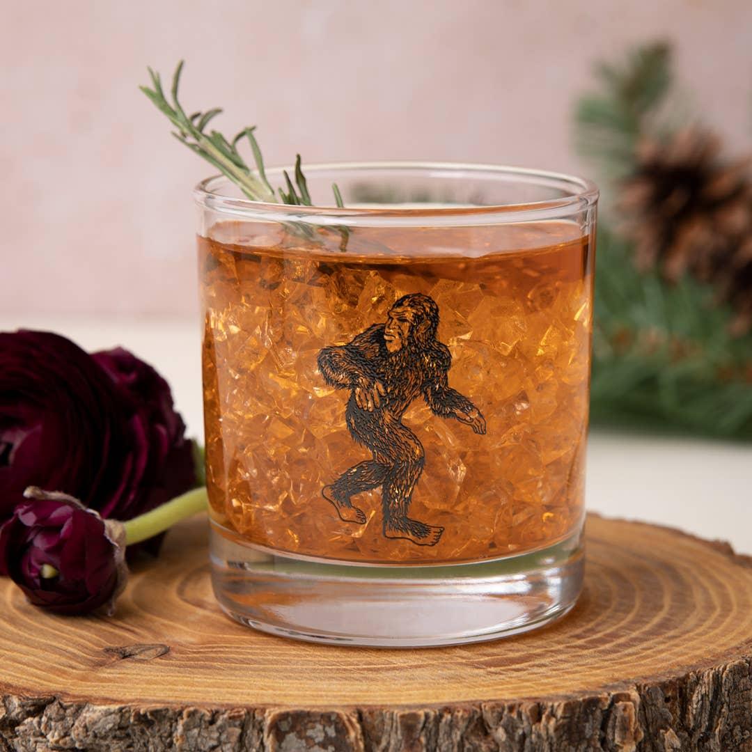 Whiskey glass with Bigfoot design, served with ice and rosemary garnish on wooden slab