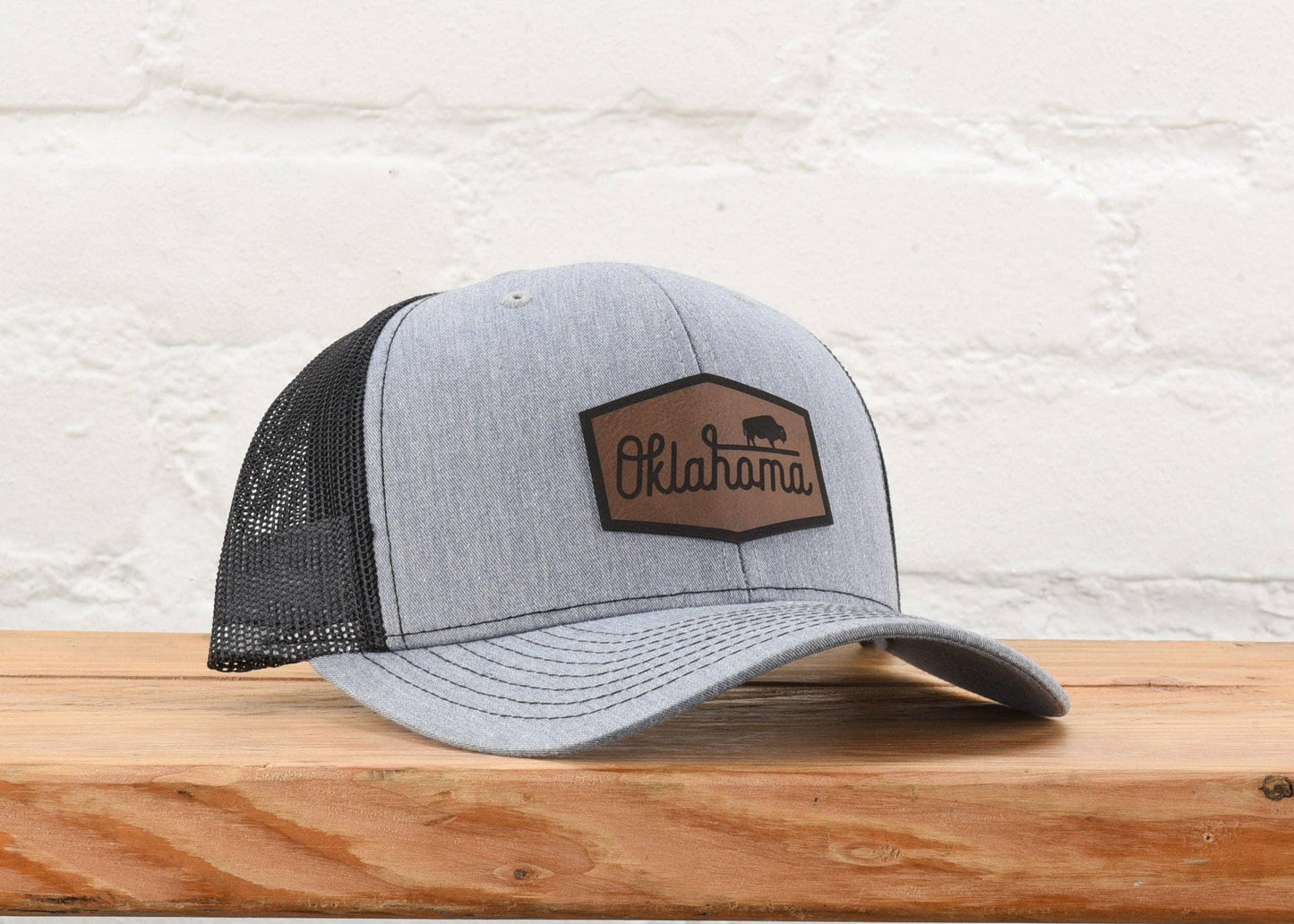 Gray trucker hat with Oklahoma bison patch, displayed on wooden surface against white brick wall