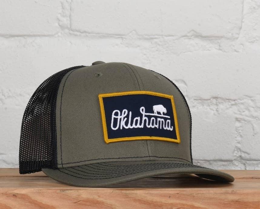 Olive green trucker hat with Oklahoma bison patch, displayed on wood against white brick