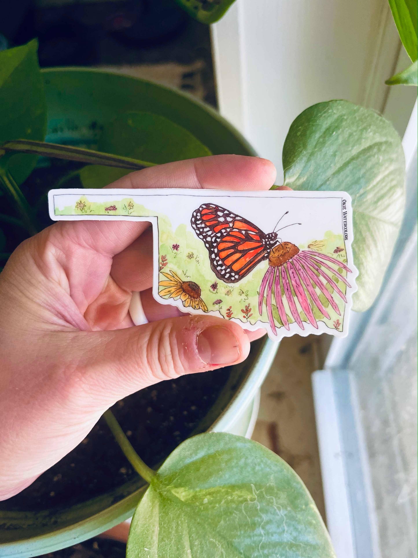 Hand holding Oklahoma-shaped sticker with monarch butterfly and wildflowers, perfect for Native art crafts.