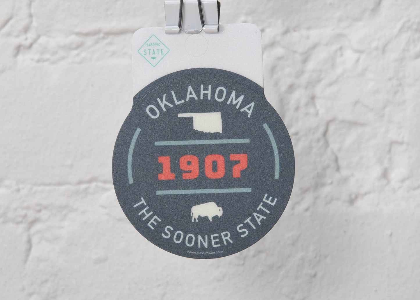 Oklahoma state sticker with 1907 and bison design, The Sooner State, hanging on white brick wall